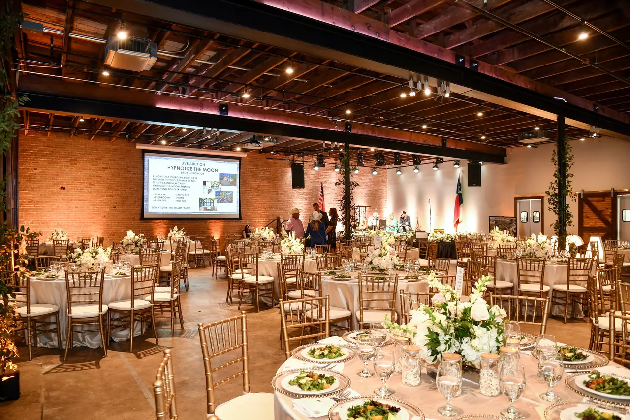 Elegant event space with round tables set for dinner, white floral arrangements, exposed brick walls, and industrial ceiling with warm lighting