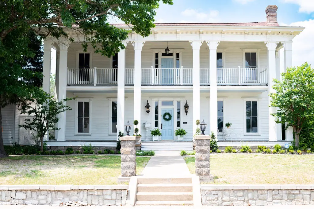 Classical white two-story mansion with columned porches, blue front door, stone pillars at entrance, and surrounding green landscaping