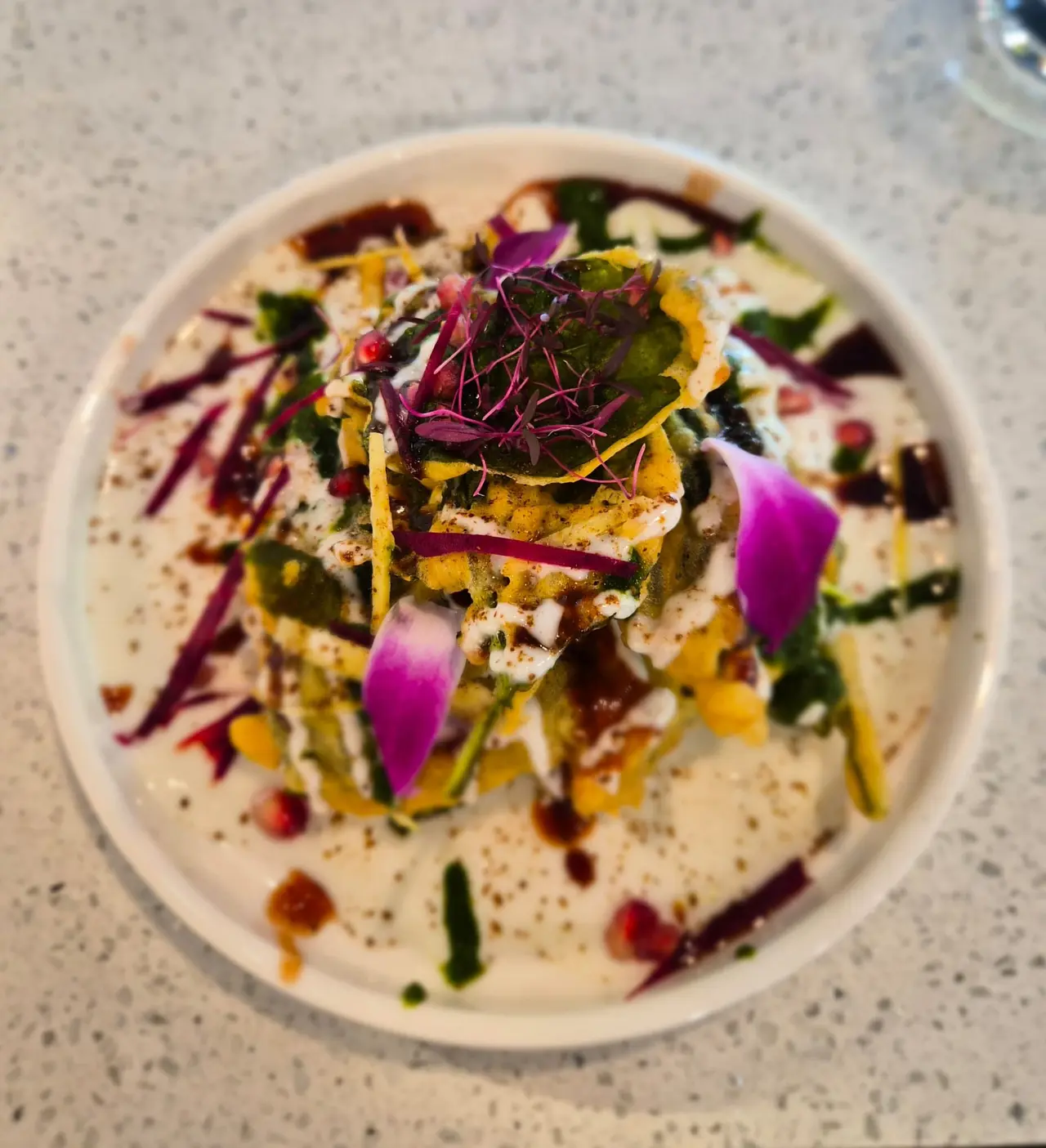 Plated dish with creamy base, topped with golden-crusted protein, purple and green microgreens, yellow garnish, and colorful edible flowers
