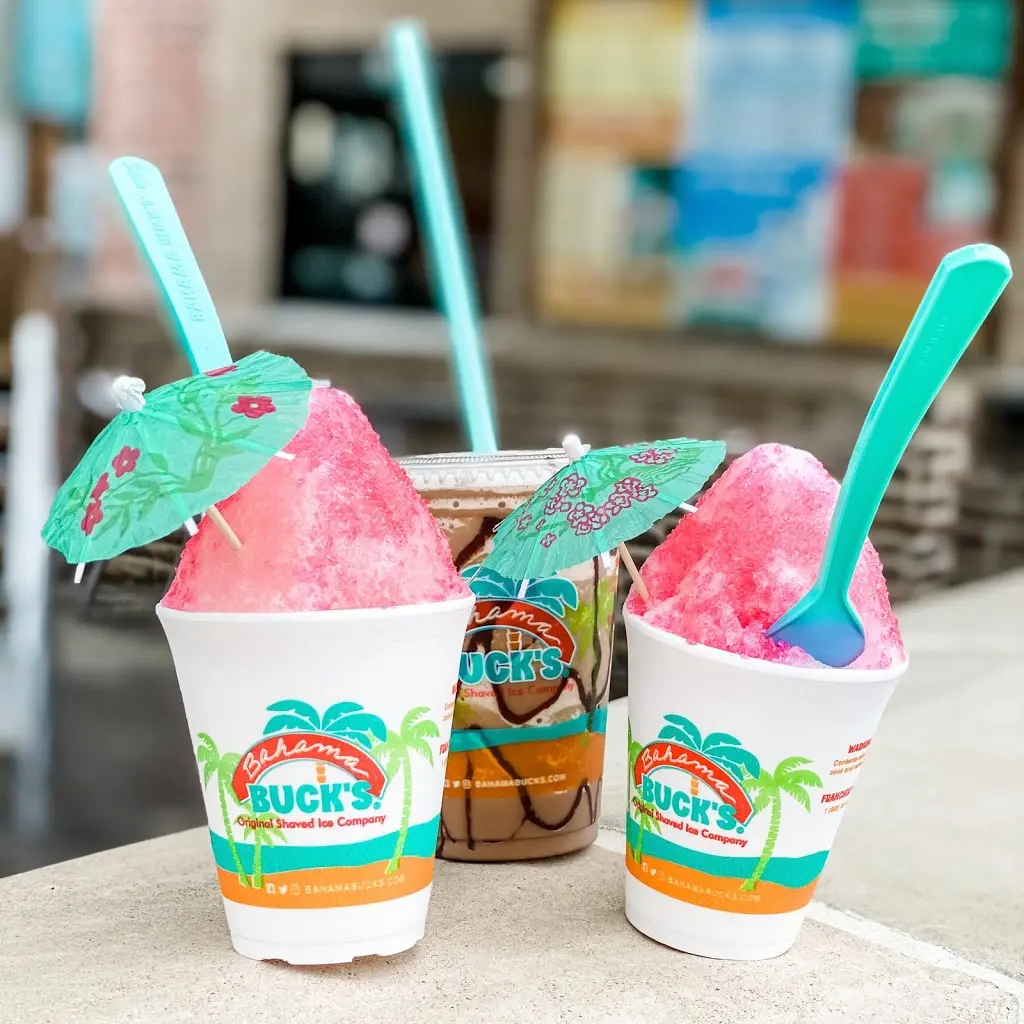 Two cups of pink shaved ice with decorative umbrellas and turquoise spoons, displayed with a jar of sauce between them