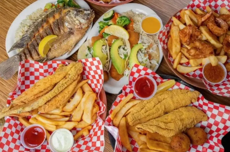 Whole fried fish with lemon, sandwiches, fried catfish, and golden fries on red and white checkered paper with sauce cups