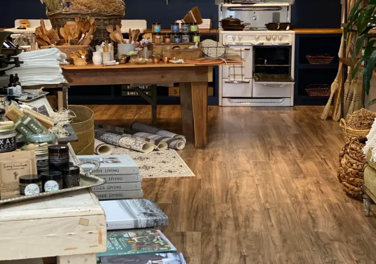 Cluttered artisan shop interior with wooden work tables, stacked linens, packaged goods, woven baskets, and kitchen items displayed throughout