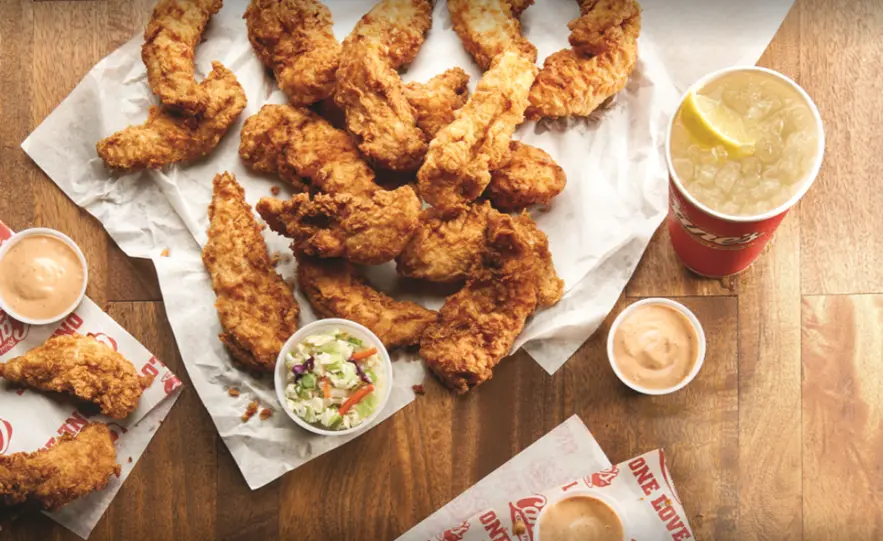 Crispy fried chicken tenders on white paper with dipping sauces and a beverage on wooden surface