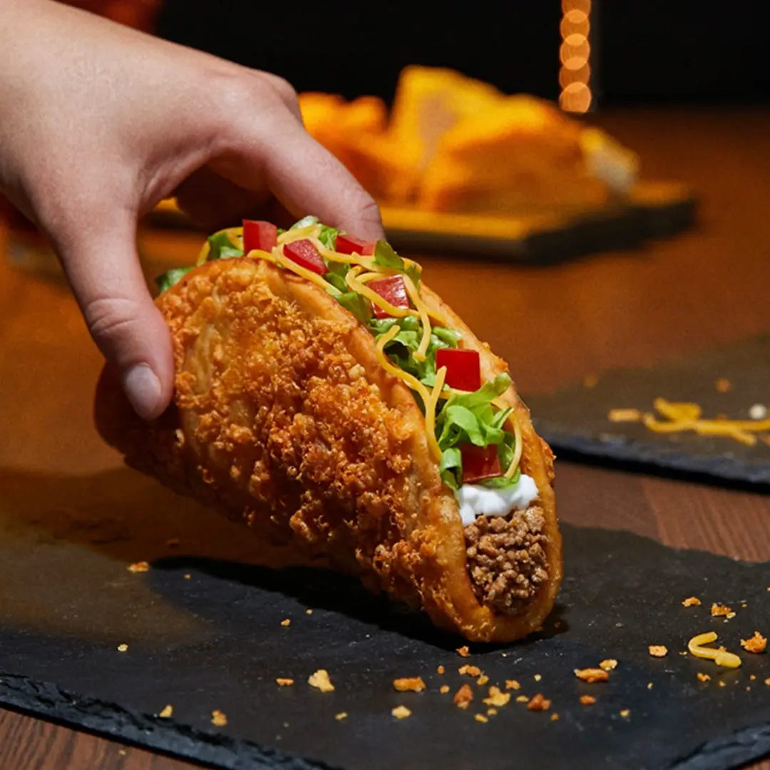 Hand holding a crispy taco topped with shredded cheese, lettuce, red peppers, and sour cream on a dark slate surface