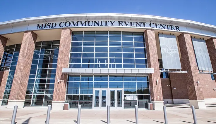 Brick and glass community event center exterior with entrance doors and metal bollards under clear blue sky