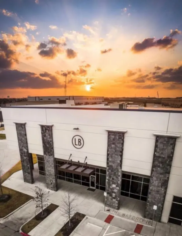 Modern white building with tall stone pillars and LB logo at sunset, surrounded by flat landscape and parked cars