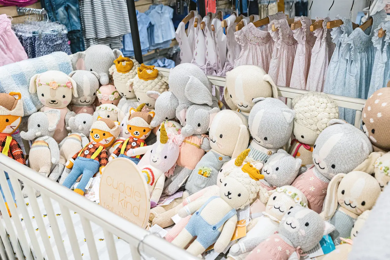 White plush toys and stuffed animals displayed in a crib with pastel-colored children's clothing hanging on racks behind