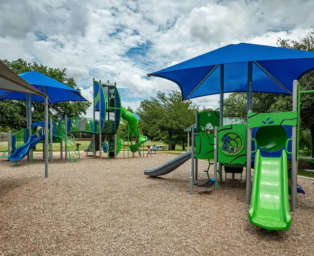 Colorful playground equipment with blue shade structures and green slides on gravel surface surrounded by trees