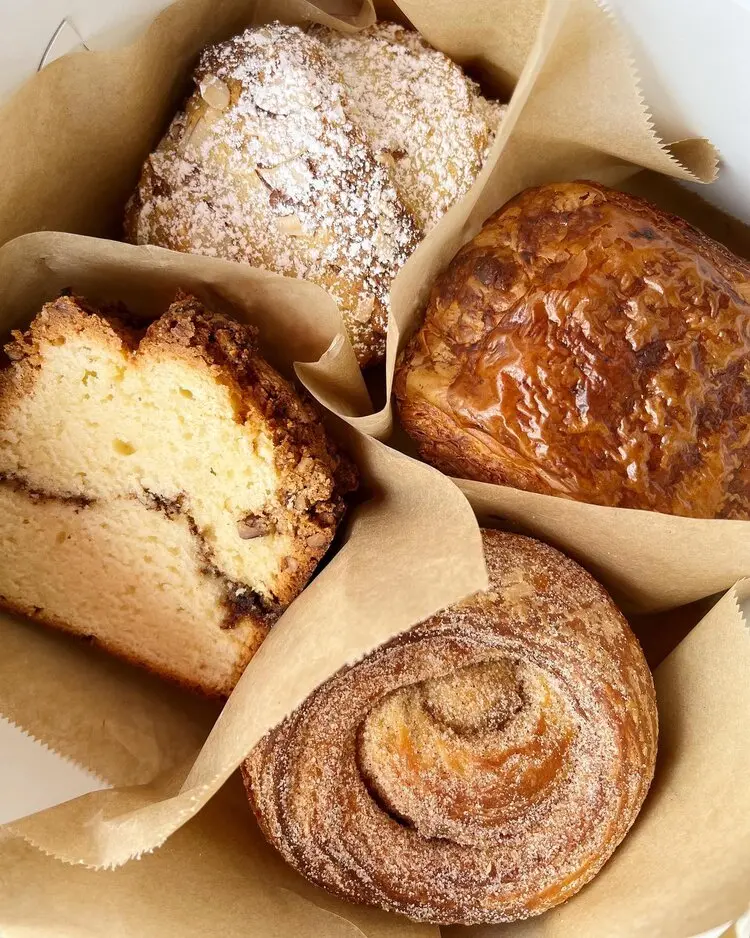 Assorted pastries and breads in parchment paper including a powdered donut, cinnamon roll, loaf cake with fruit, and dense brown bread