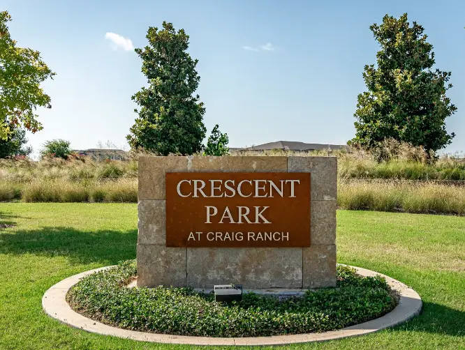 Brown stone monument sign reading Crescent Park at Craig Ranch, surrounded by landscaping with green grass and trees in background