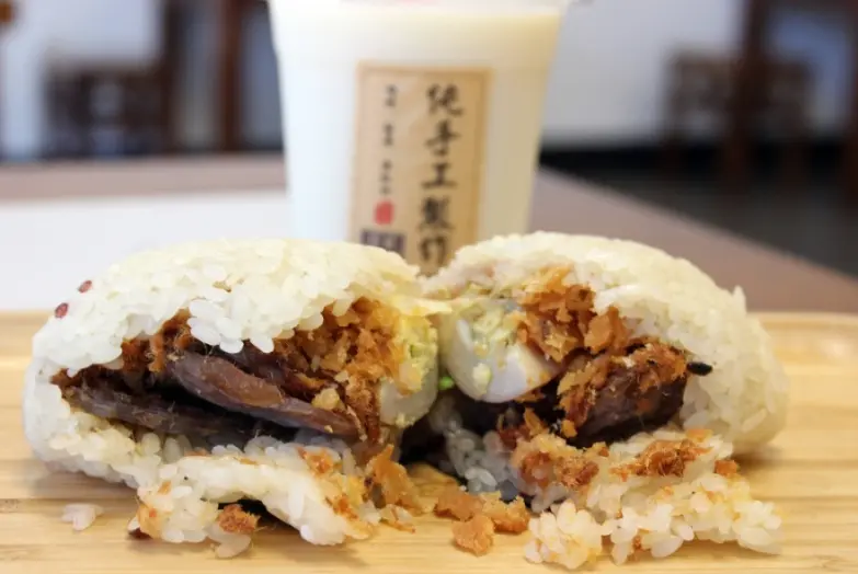 Rice roll sandwich filled with fried shallots, meat, and egg on wooden board with beverage in background