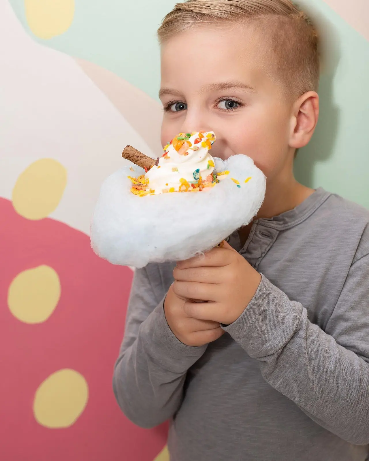 Young child eating cotton candy ice cream with whipped cream and colorful sprinkles against colorful polka dot background