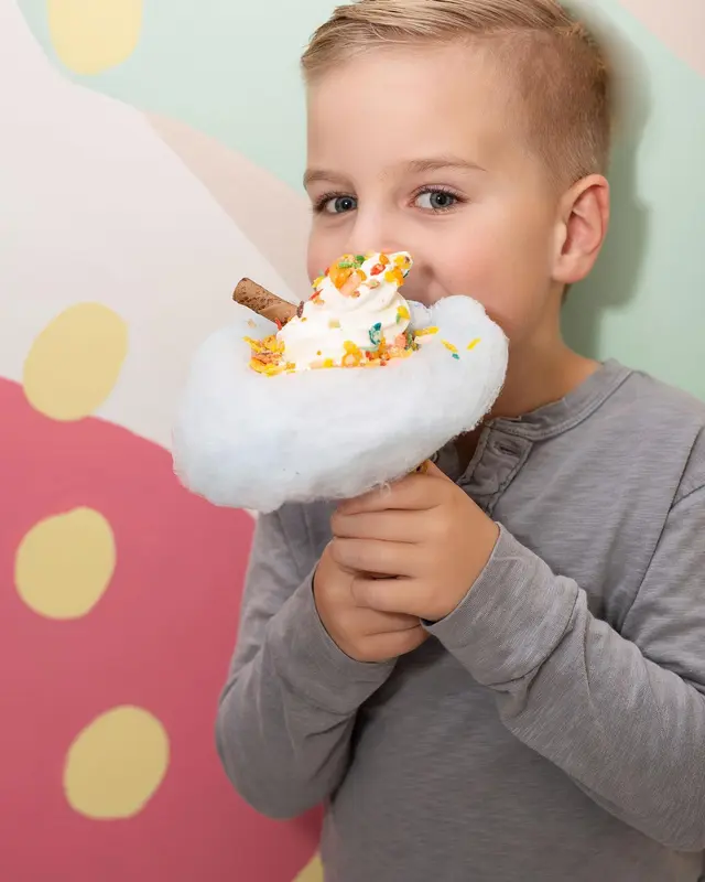 Young child eating cotton candy ice cream with whipped cream and colorful sprinkles against colorful polka dot background
