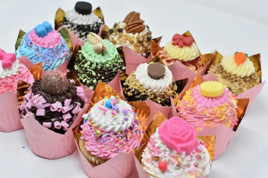 Colorful frosted cupcakes in pink paper wrappers with sprinkles, candy toppings, and gold leaf accents arranged on white background