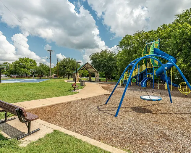Blue playground structure with slide and climbing elements on mulch ground, surrounded by green lawn and trees under cloudy sky