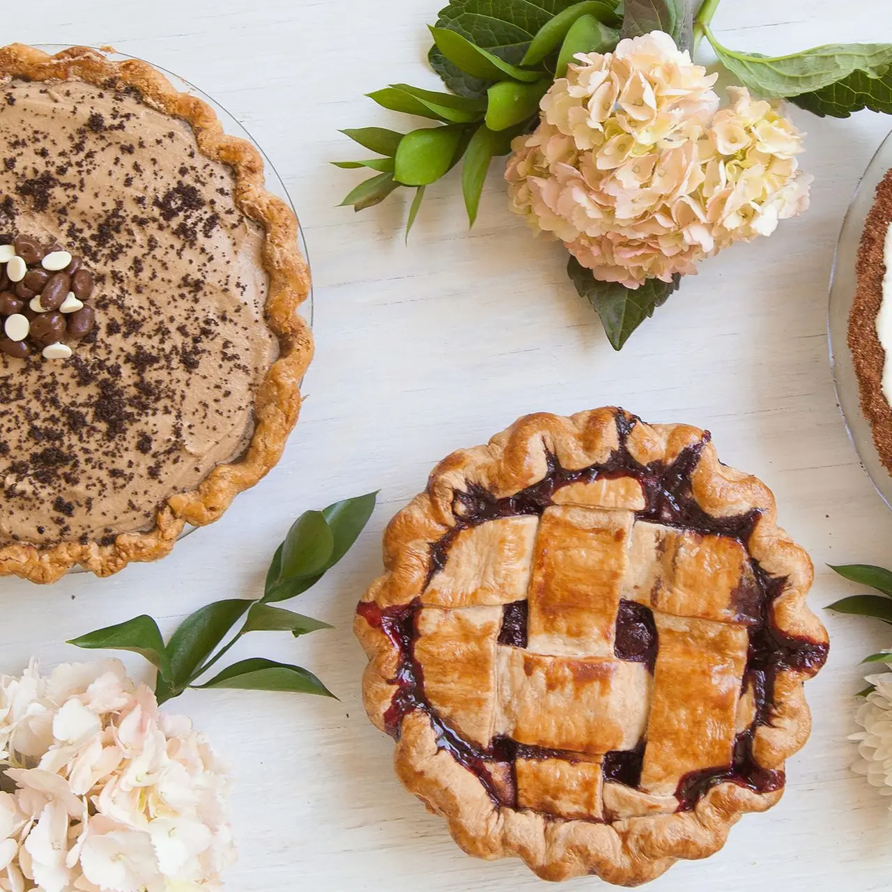 Assorted handmade pies with crimped edges and lattice crust, arranged with fresh flowers and green foliage on white surface