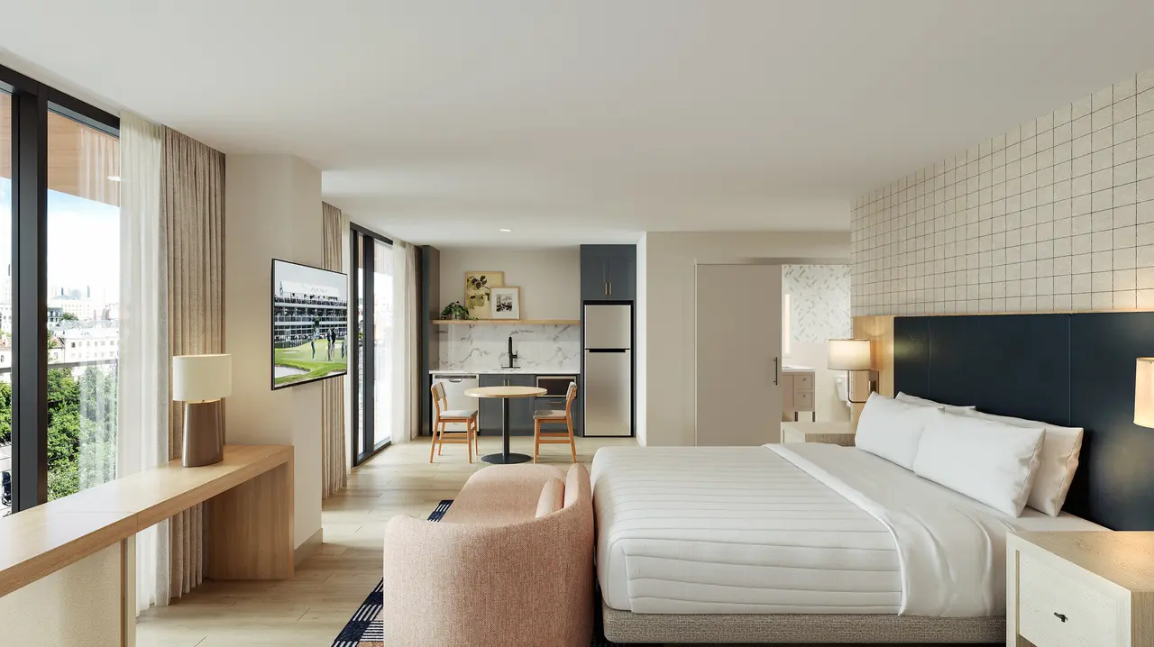 Modern hotel room with bed, lounge chair, desk area, kitchenette, and floor-to-ceiling windows overlooking city street
