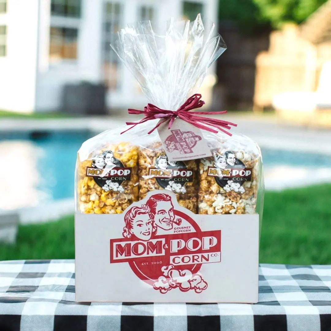 Three bags of gourmet popcorn with branded labels tied with burgundy ribbon, displayed on checkered tablecloth with pool and house in background