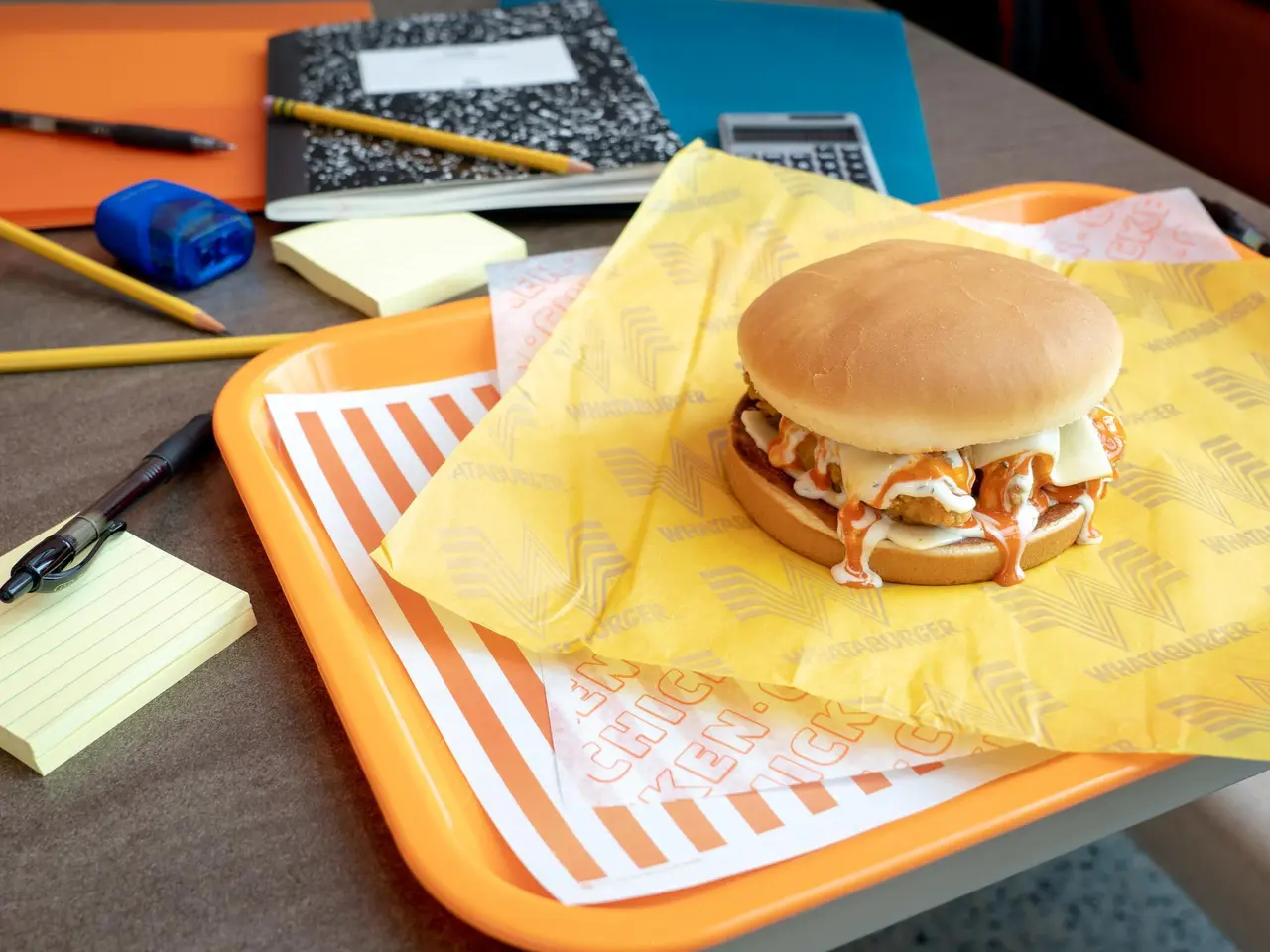 Pulled pork sandwich on yellow paper wrapper atop orange striped tray on desk with school supplies and notebook