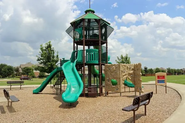 Tall playground structure with green slide and climbing features on sandy ground, surrounded by benches and green grass field
