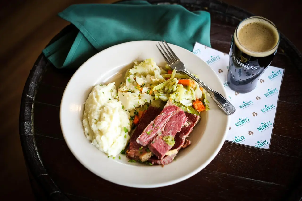 Plate of corned beef, mashed potatoes, and mixed vegetables with fork, napkin, and pint glass on dark table