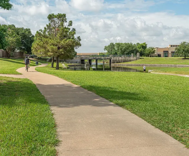 Paved pathway through green lawn with trees, bridge over water, and sports fields in background under blue sky