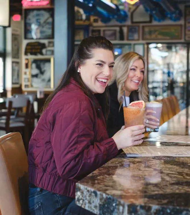Two women smiling at a bar counter, each holding colorful cocktails, with restaurant interior and signage visible behind them