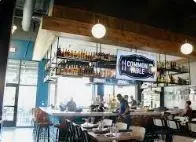 Restaurant interior with wooden ceiling, pendant lights, shelving with bottles, and diners seated at tables