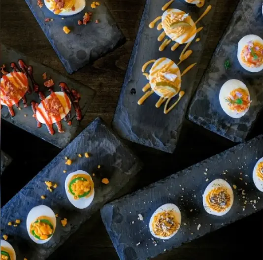 Small bowls of deviled eggs with various toppings arranged on dark slate boards, overhead view