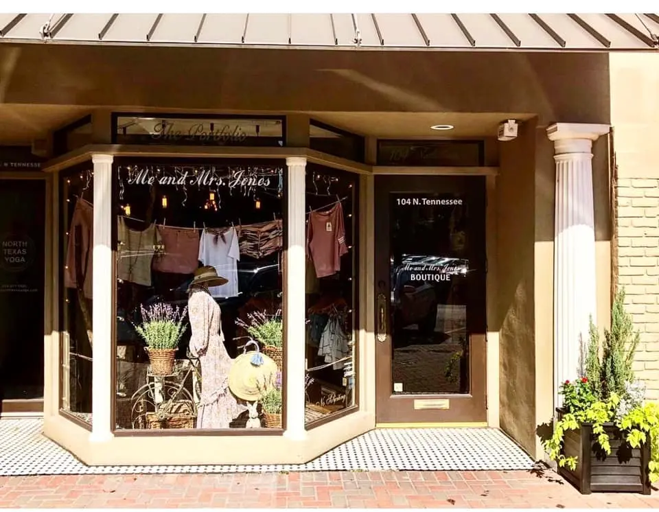 Storefront with angled glass windows displaying clothing, accessories, and floral arrangements in earth tones, brick facade with potted plants flanking the