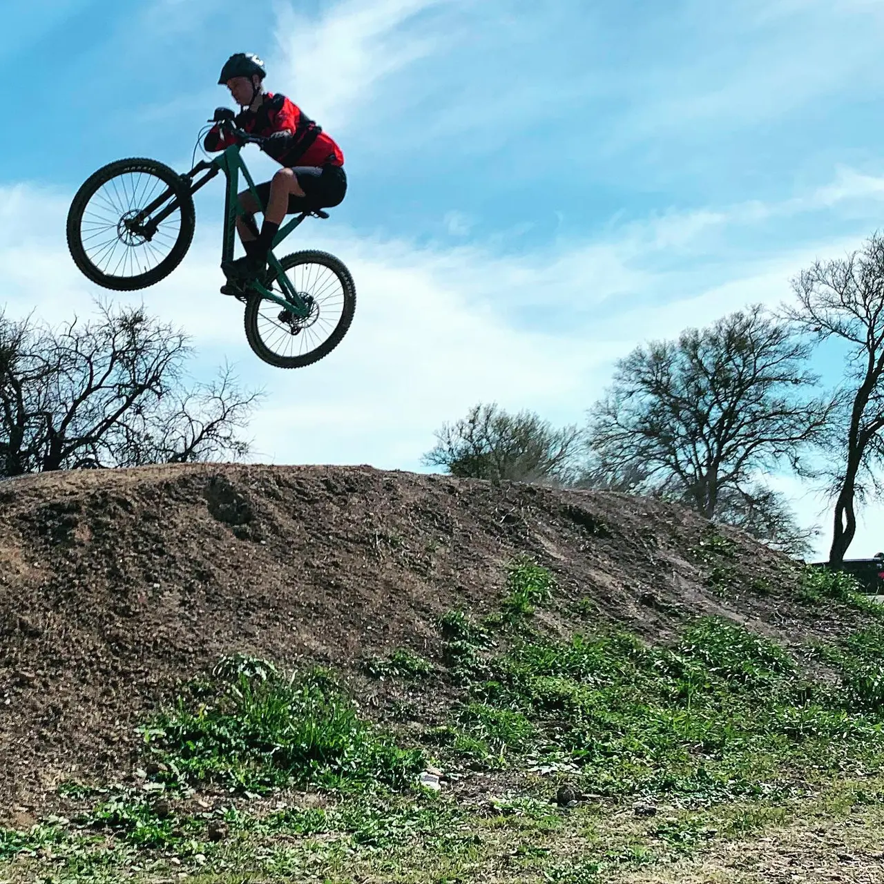 Mountain biker performing aerial trick off dirt jump against blue sky and bare trees