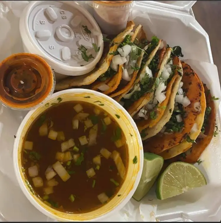 Tacos with lettuce and cheese on white plate, served with broth soup, salsa, and lime wedges