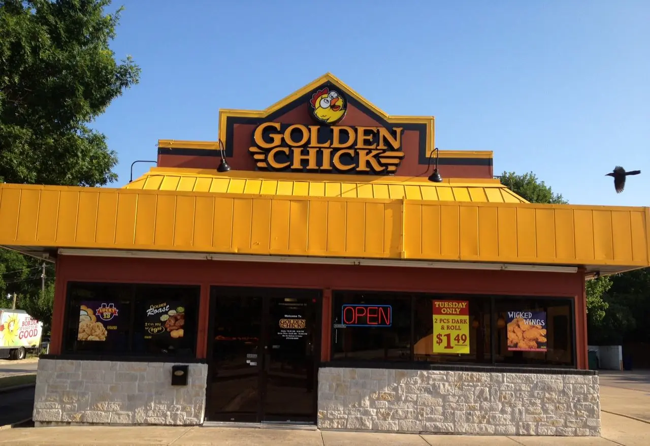 Red and yellow storefront with golden chicken sign, stone base, and open neon sign, blue sky above