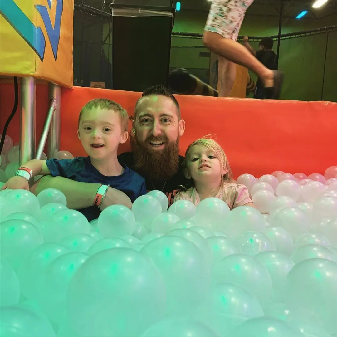 Adult and two young children in a ball pit with glowing turquoise balls, red wall backdrop, indoor play facility
