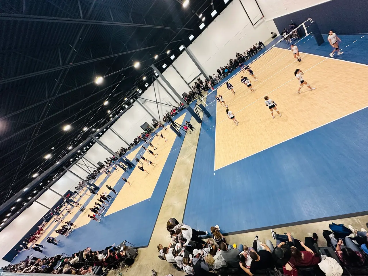 Indoor volleyball court with spectators in bleachers watching players compete on blue and sand-colored court under bright overhead lighting