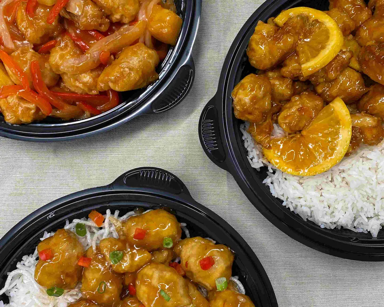 Three black takeout containers with glazed meatballs and shrimp over white rice, garnished with lemon slices and red peppers