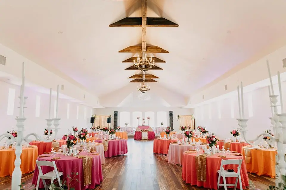 Bright event space with wooden ceiling fixtures, round tables dressed in coral and magenta linens, white chairs, and floral centerpieces leading to a decorated