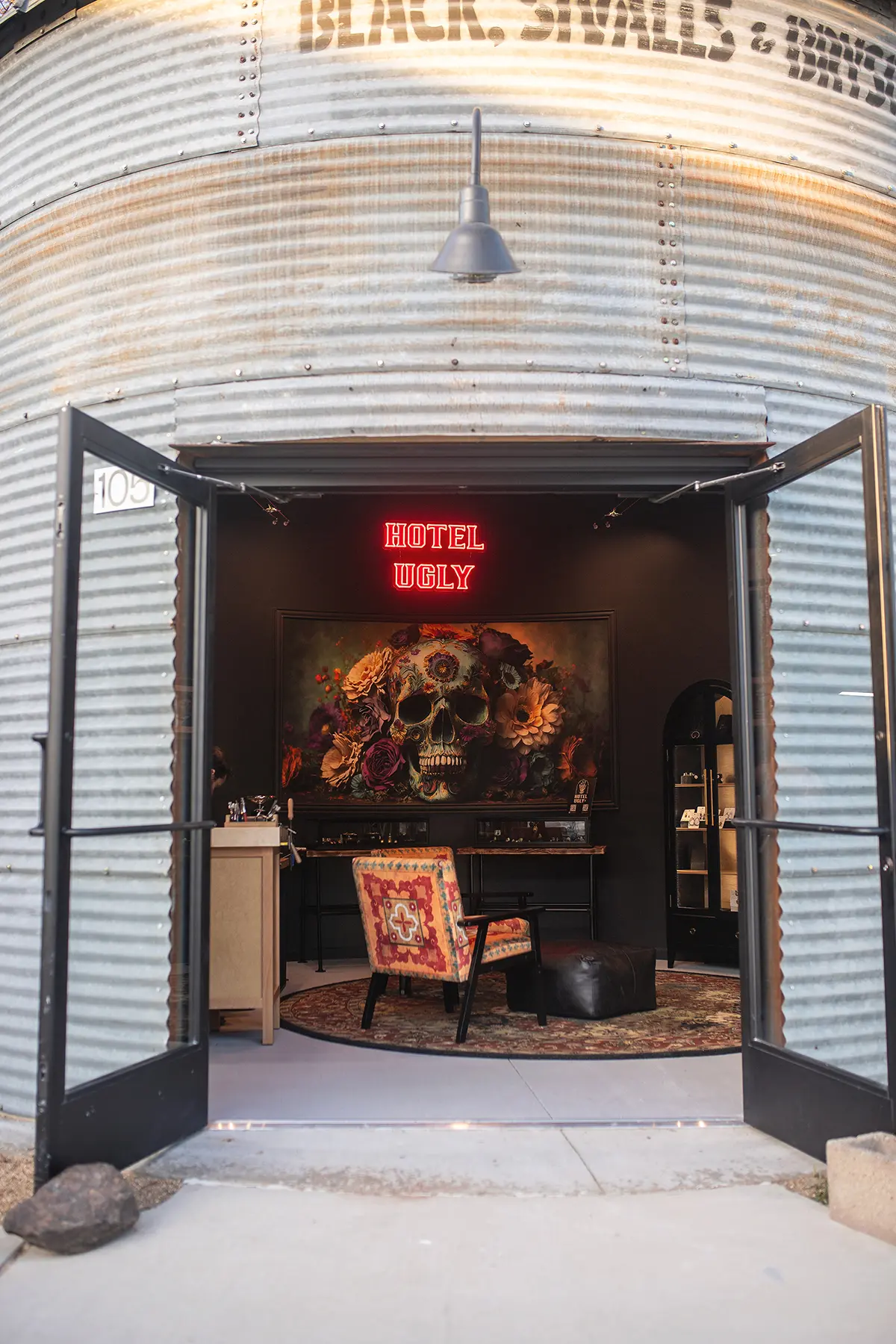 Metal storefront with open doors revealing neon sign, skull artwork, jewelry display, and vintage chair inside