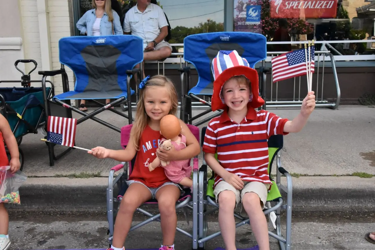 Two kids sitting in lawn chairs at a parade waving American flags