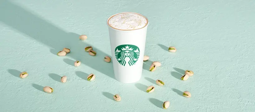 White paper cup with whipped cream top surrounded by scattered coffee beans on mint green surface