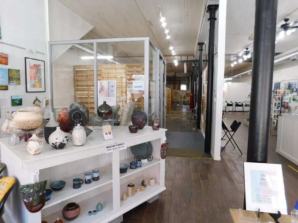 Gallery interior with white display shelves holding ceramic bowls and vessels, glass-fronted exhibition case, framed artwork on walls, and wooden flooring