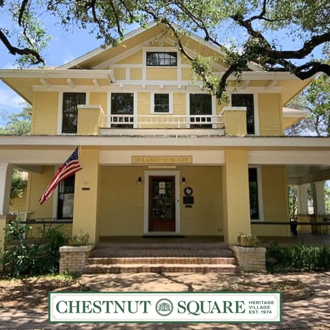Yellow two-story historic house with white trim, American flag, and front porch with columns, surrounded by large oak trees