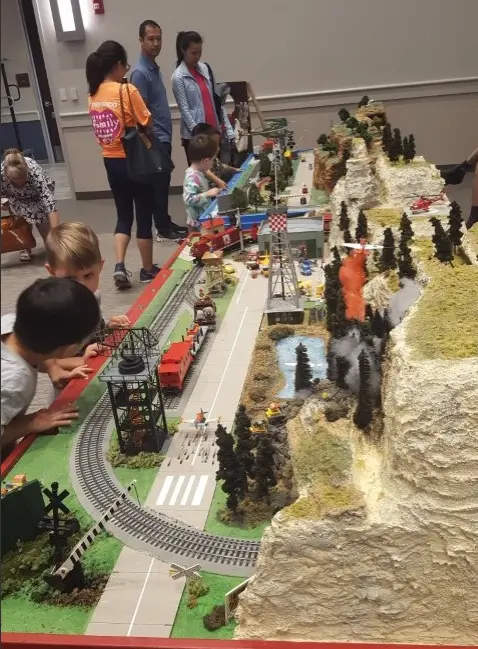 Children and adults viewing an elaborate model train layout with miniature trees, tracks, and scenery on display tables