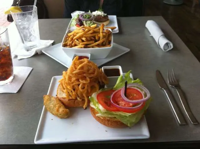 Plated burger with crispy onion strings, lettuce and tomato, served with curly fries and onion rings, alongside drinks and napkins on a dark table