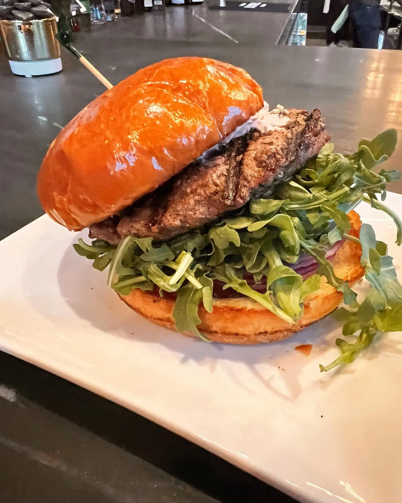 Burger with orange bun, grilled meat patty, and mixed greens on white plate at restaurant counter