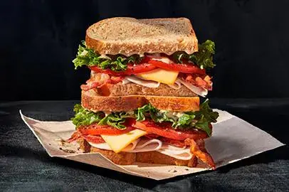Stacked sandwich with toasted bread, lettuce, tomato, cheese, and cured meat on parchment paper