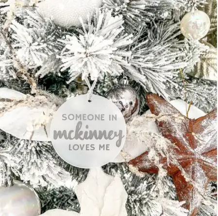 Frost-covered evergreen branches with red and silver ornaments and a white circular ornament reading "Someone in McKinney loves me