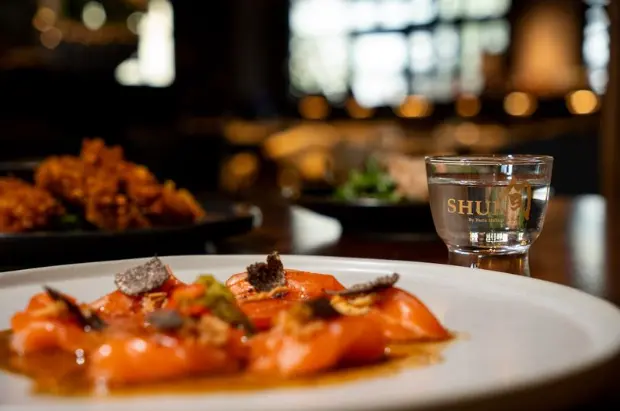 Plated orange-glazed dish with garnish and a glass of liquor on a restaurant table with blurred warm lighting in background