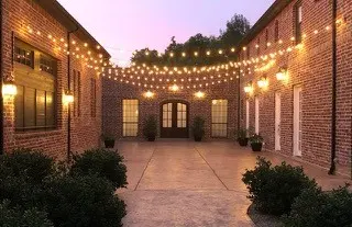 Brick courtyard with string lights overhead, illuminated sconces on walls, potted plants, and arched doorways at dusk