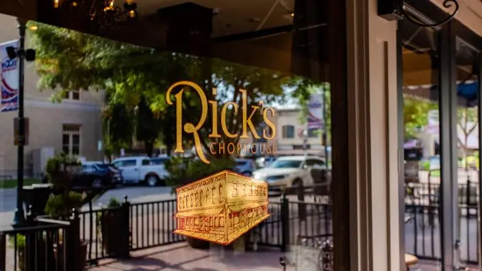 Gold lettering and logo on restaurant window with street view and outdoor seating visible beyond glass doors